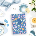26/27 Weekly Monthly Planner on a white tabletop beside a candle, lemon water, flowers, and blue accessories.