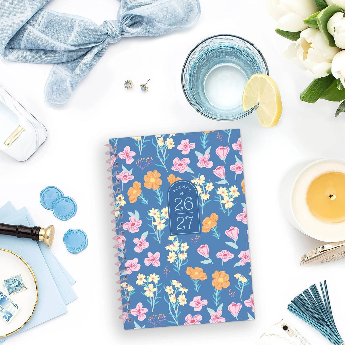 26/27 Weekly Monthly Planner on a white tabletop beside a candle, lemon water, flowers, and blue accessories.