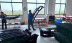 Supplier workshop showing workers handling automotive body components at worktables.
