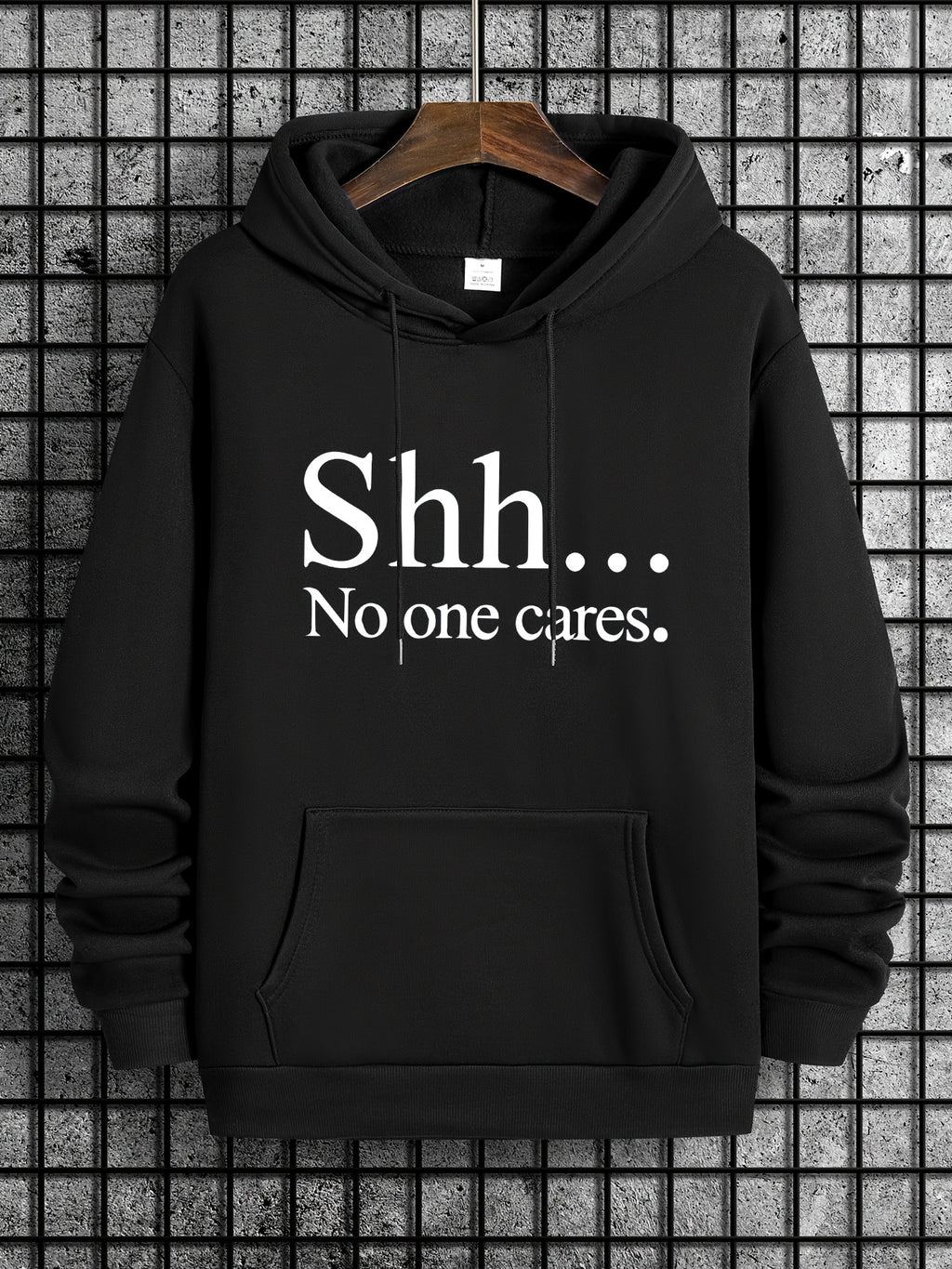 Black SHH No One Cares hoodie with white text, front kangaroo pocket on wooden hanger against tiled wall