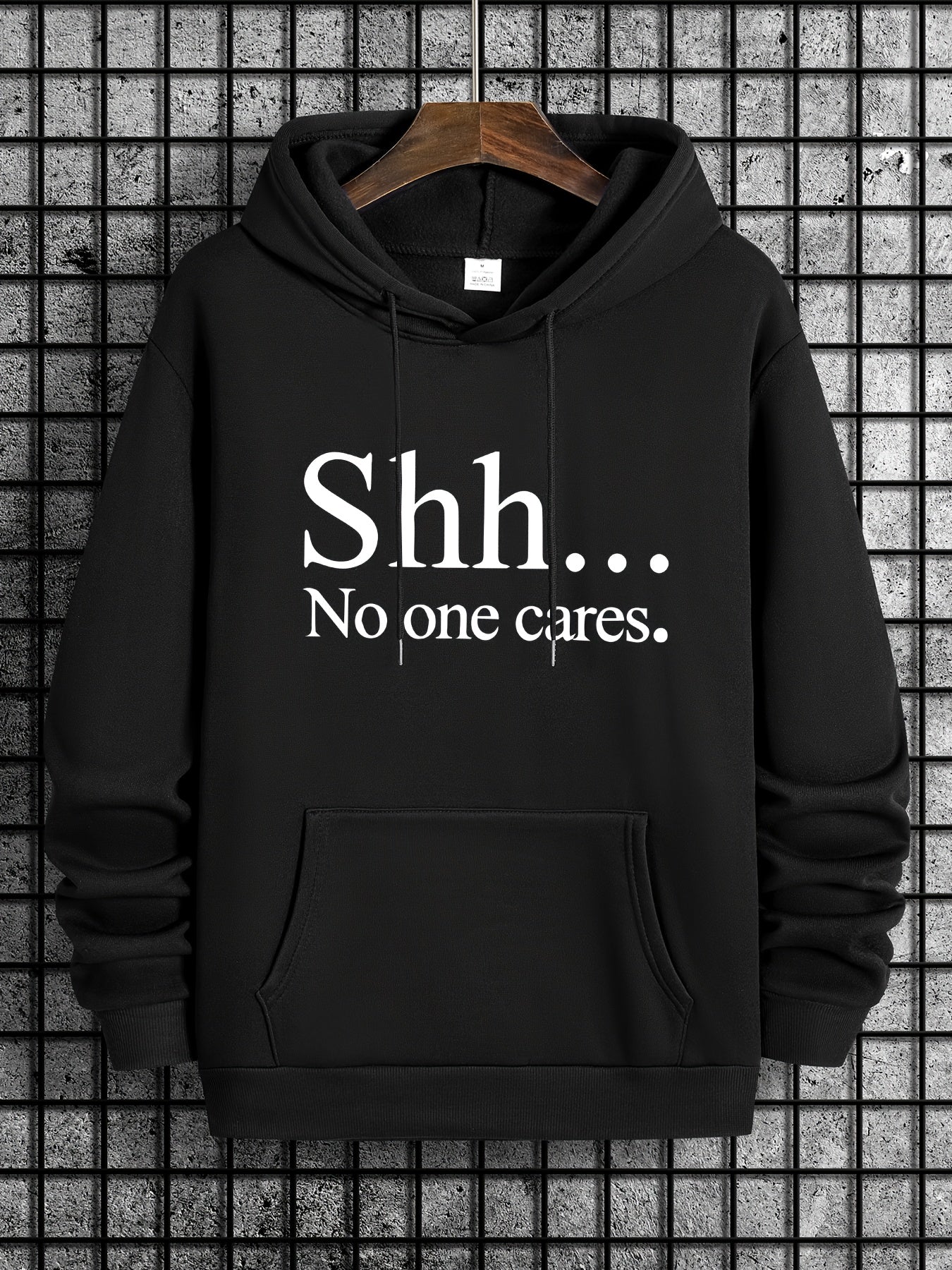 Black SHH No One Cares hoodie with white text, front kangaroo pocket on wooden hanger against tiled wall