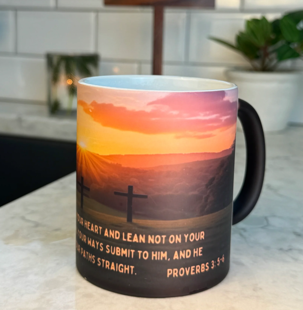 FaithGlow Mug featuring Proverbs 3:5-6 with sunset mountain scene and cross silhouette on black ceramic, heat-reactive design