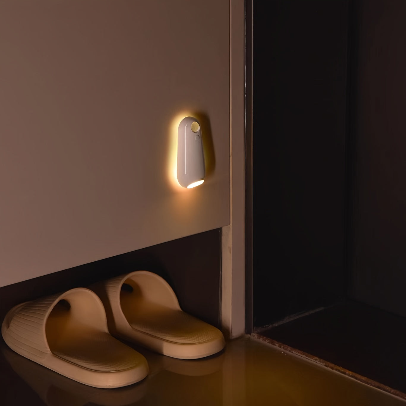 GlowStep Motion Sensor Light mounted on wall, emitting warm yellow glow above house slippers in dark hallway setting