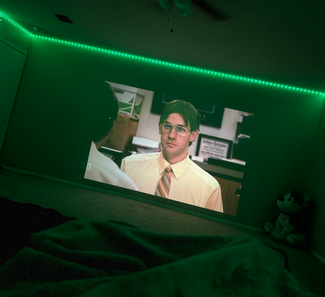 HighPeak INFINITY MiniProjector displaying a movie scene in a dark room with green LED ambient lighting and wall decoration