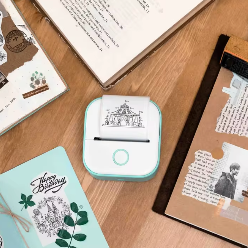 HighPeak MiniPrinter in mint green showing carousel design print, displayed on wooden desk with birthday card and study materials