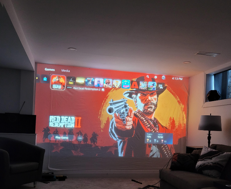 HighPeak MiniProjector SMART displaying Red Dead Redemption 2 game screen on wall in dim living room with cozy sofa setup