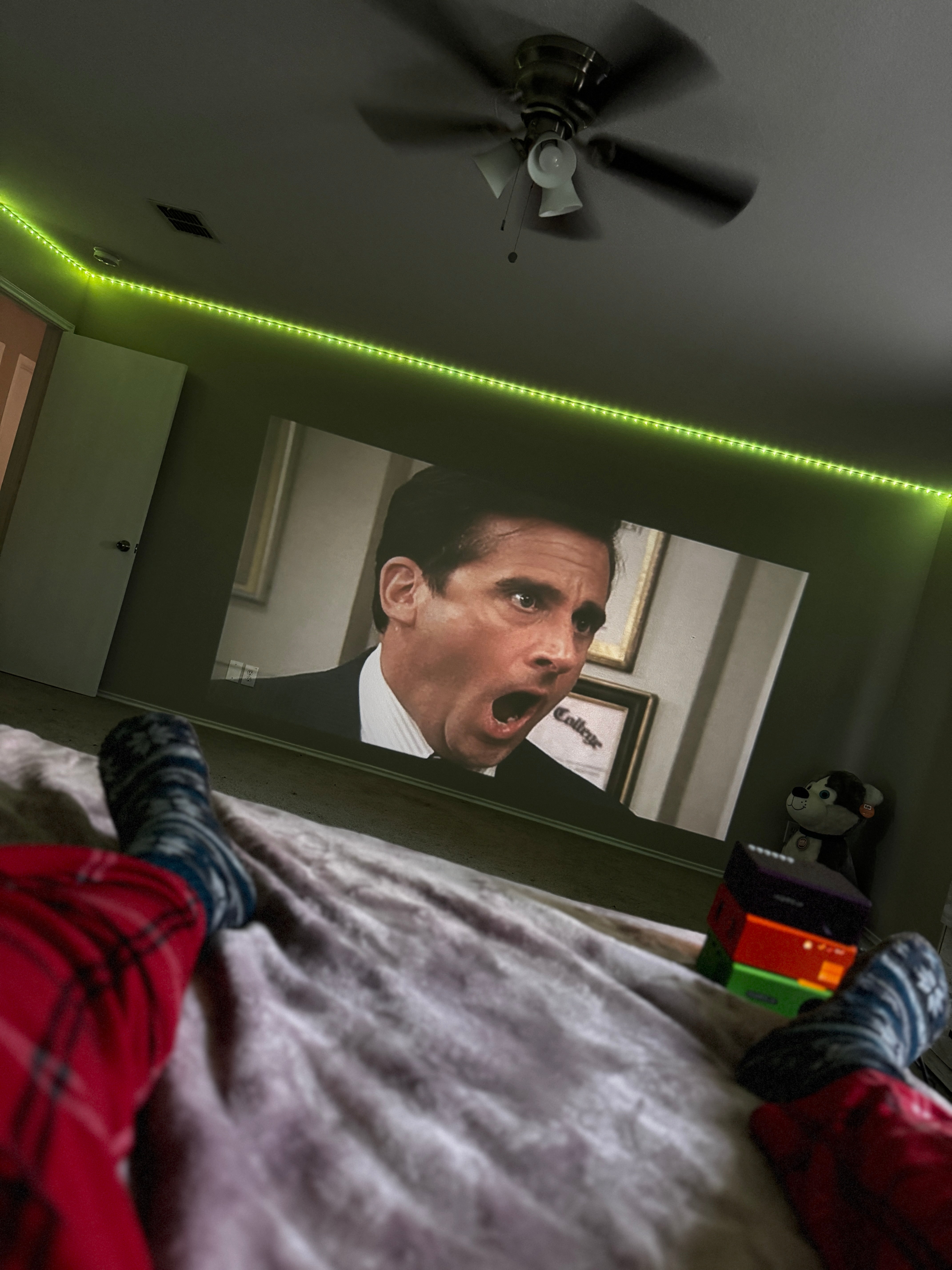 HighPeak MiniProjector SMART displaying movie on wall with green LED ambiance lights and ceiling fan in cozy bedroom setup