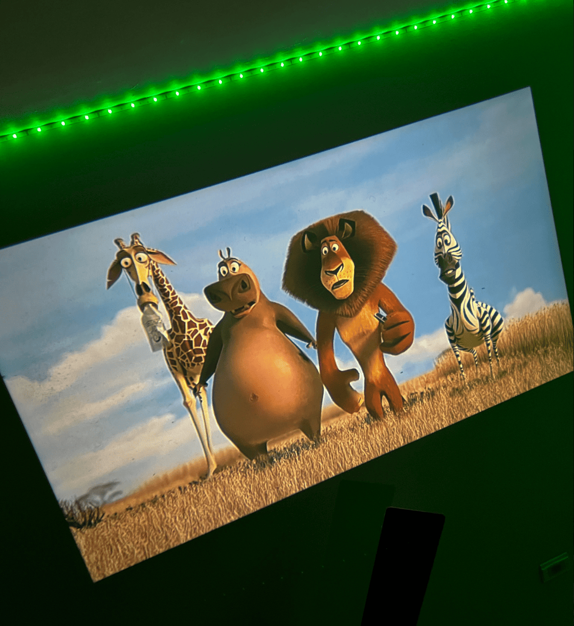 HighPeak MiniProjector™ projecting a vibrant animated movie scene on a wall with green ambient LED lighting.