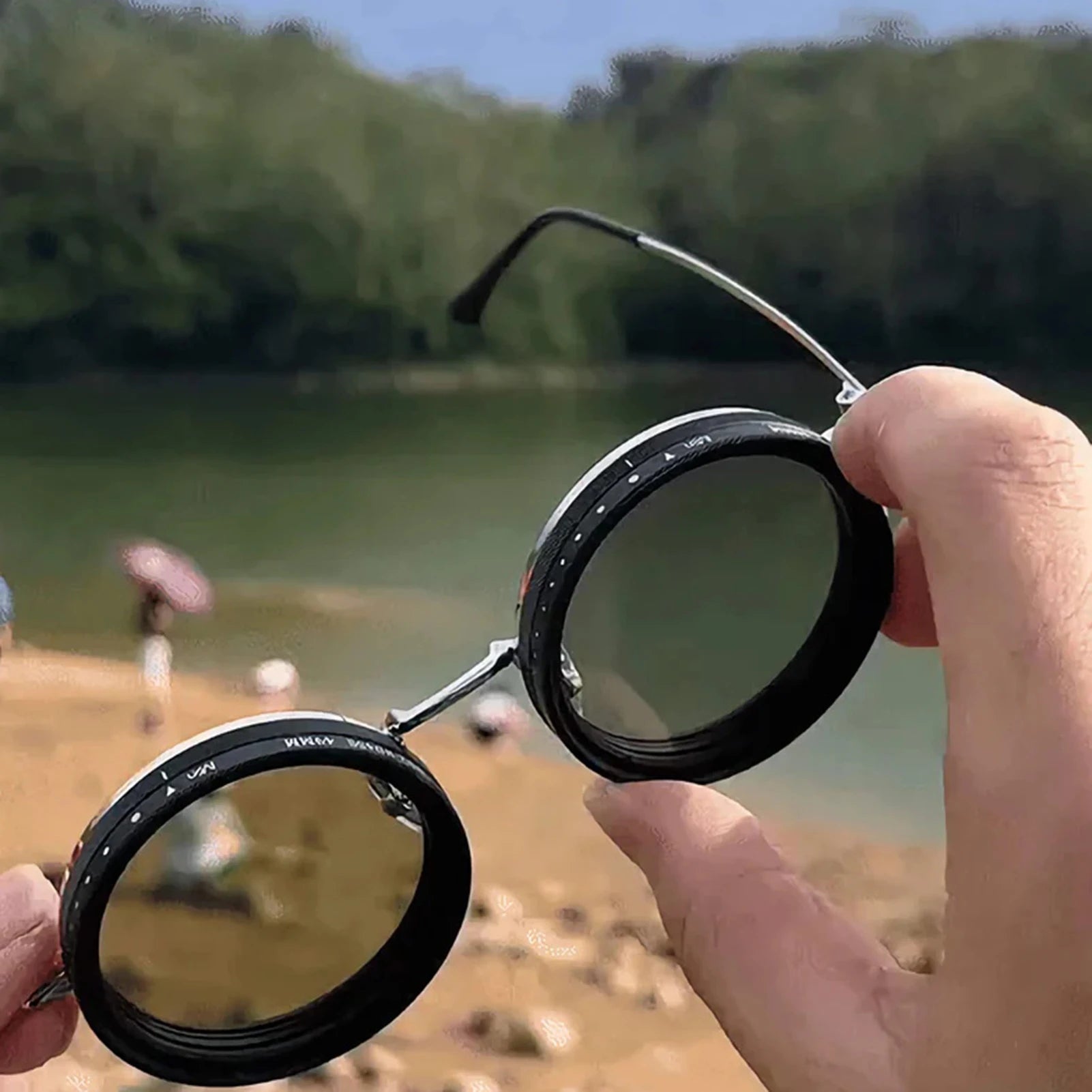 HighPeak ShiftLens™ round black adjustable sunglasses held up against lakeside nature background showing adaptive lens system