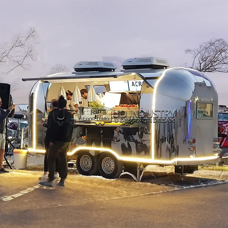 HighPeak Airstream Food Trailer with polished stainless steel exterior and illuminated serving window at an outdoor evening event