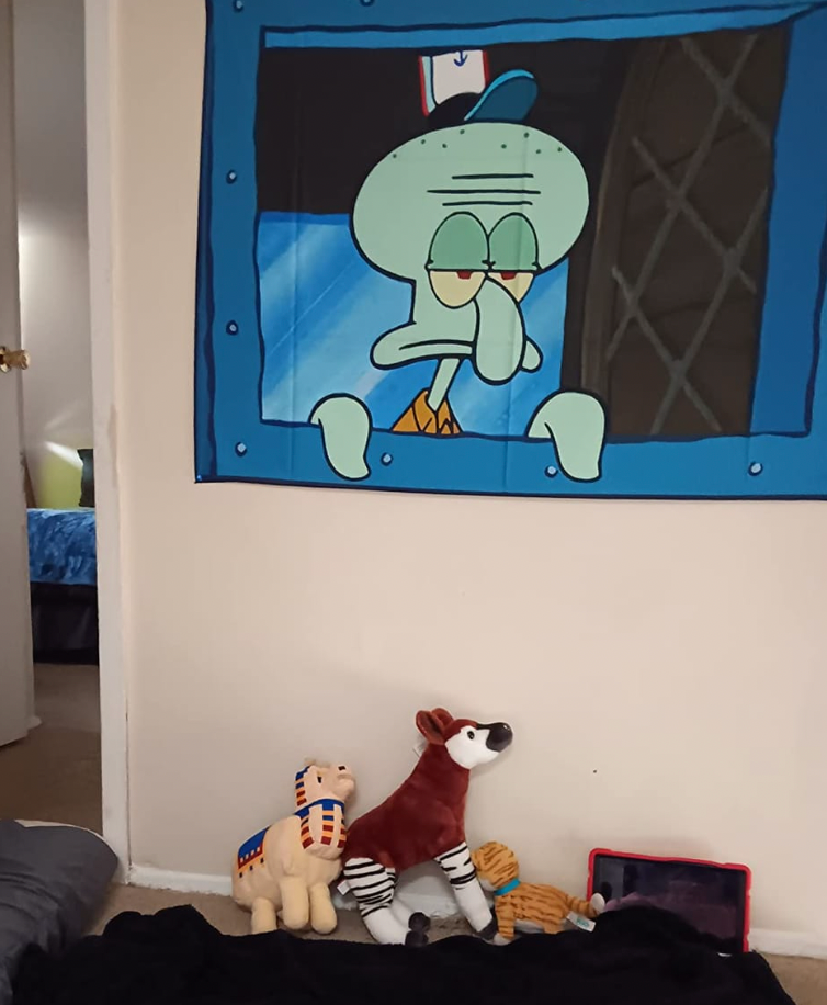 Tentacle Vibes wall tapestry featuring Squidward at window with grumpy expression, displayed above decorative plush toys