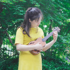 Acoustic Four-String Ukulele player in a yellow shirt holding the warm brown version among green trees.