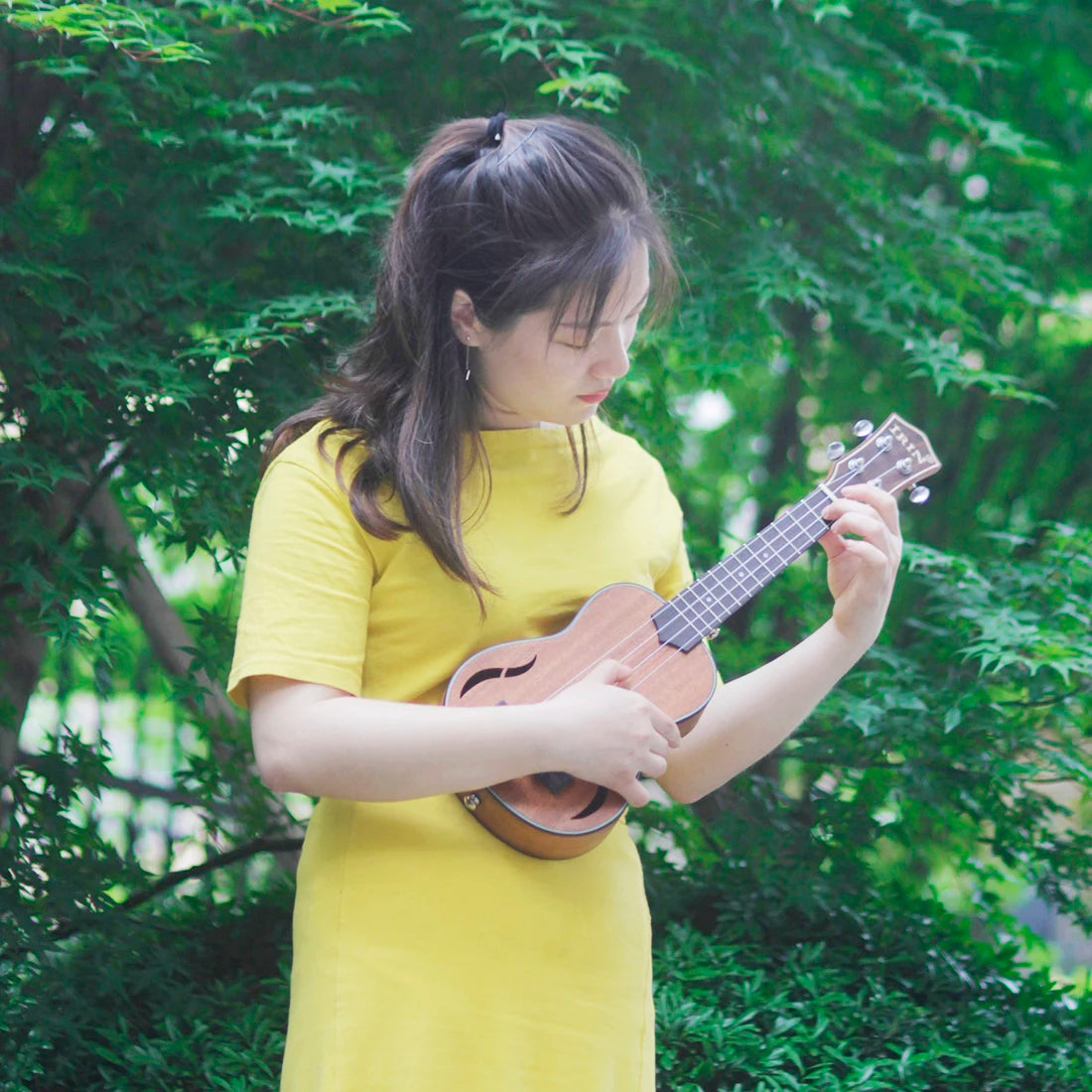 Acoustic Four-String Ukulele player in a yellow shirt holding the warm brown version among green trees.