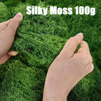 Artificial Decorative Moss held between two hands to show fine green strands, with a 100 g label.