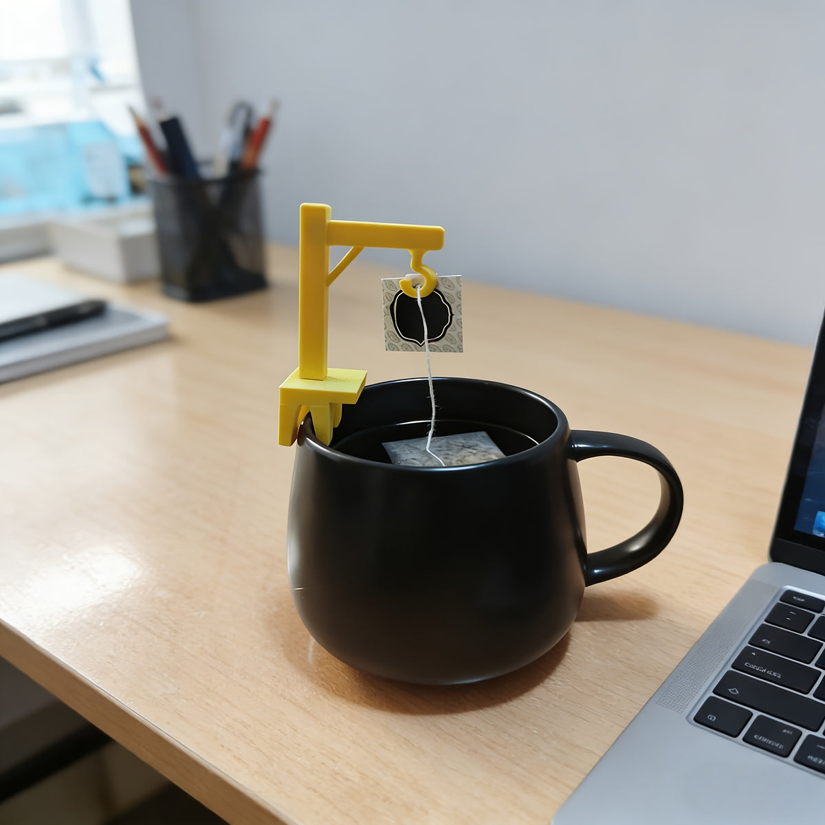 Crane Tea Bag Holder holding a tea bag string on a black mug beside a laptop on a wooden desk.