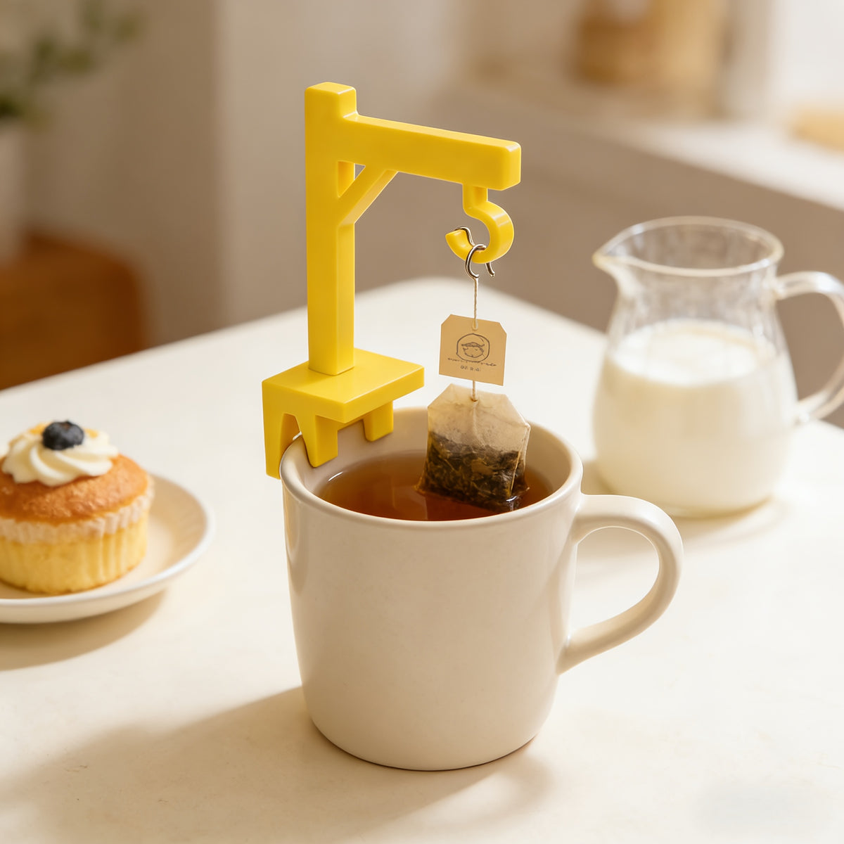 Crane Tea Bag Holder clipped to a cream mug with a tea bag hanging above the drink beside a cupcake.