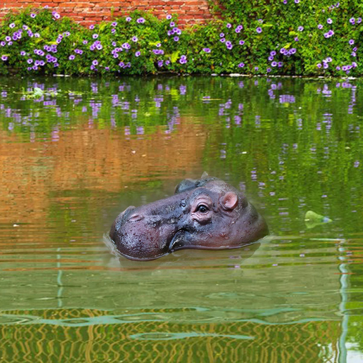 Floating Hippo Pond Decor left-facing hippo head floating in a garden pond with flowers and a brick wall behind it.