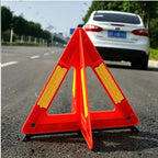 Foldable Warning Triangle close-up on asphalt behind a white parked car, showing its reflective side panels.