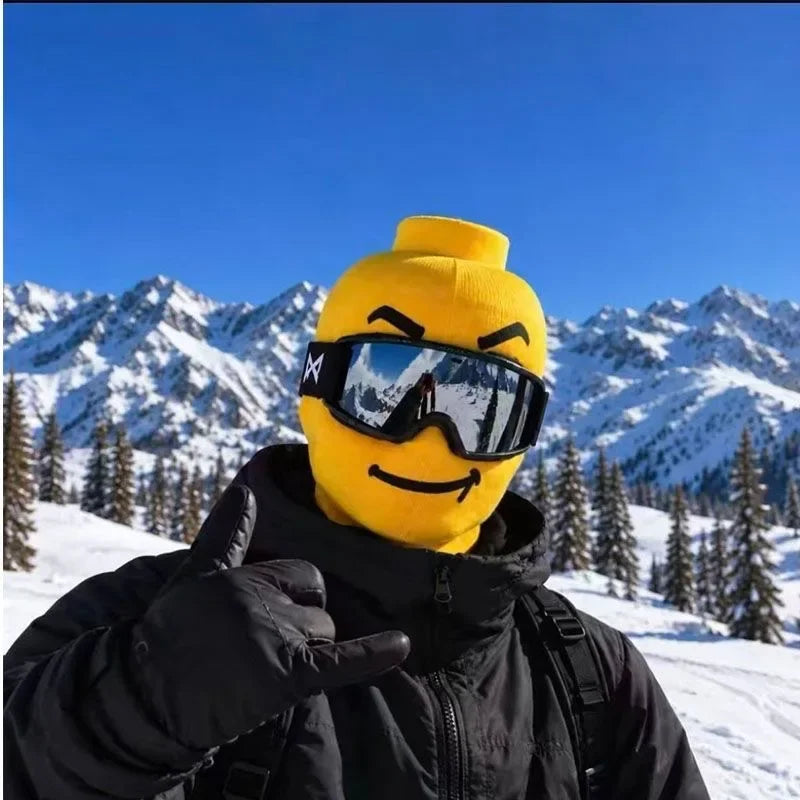Funny Brickhead Ski Mask with a simple toothless smile, worn with goggles and a black jacket in the mountains