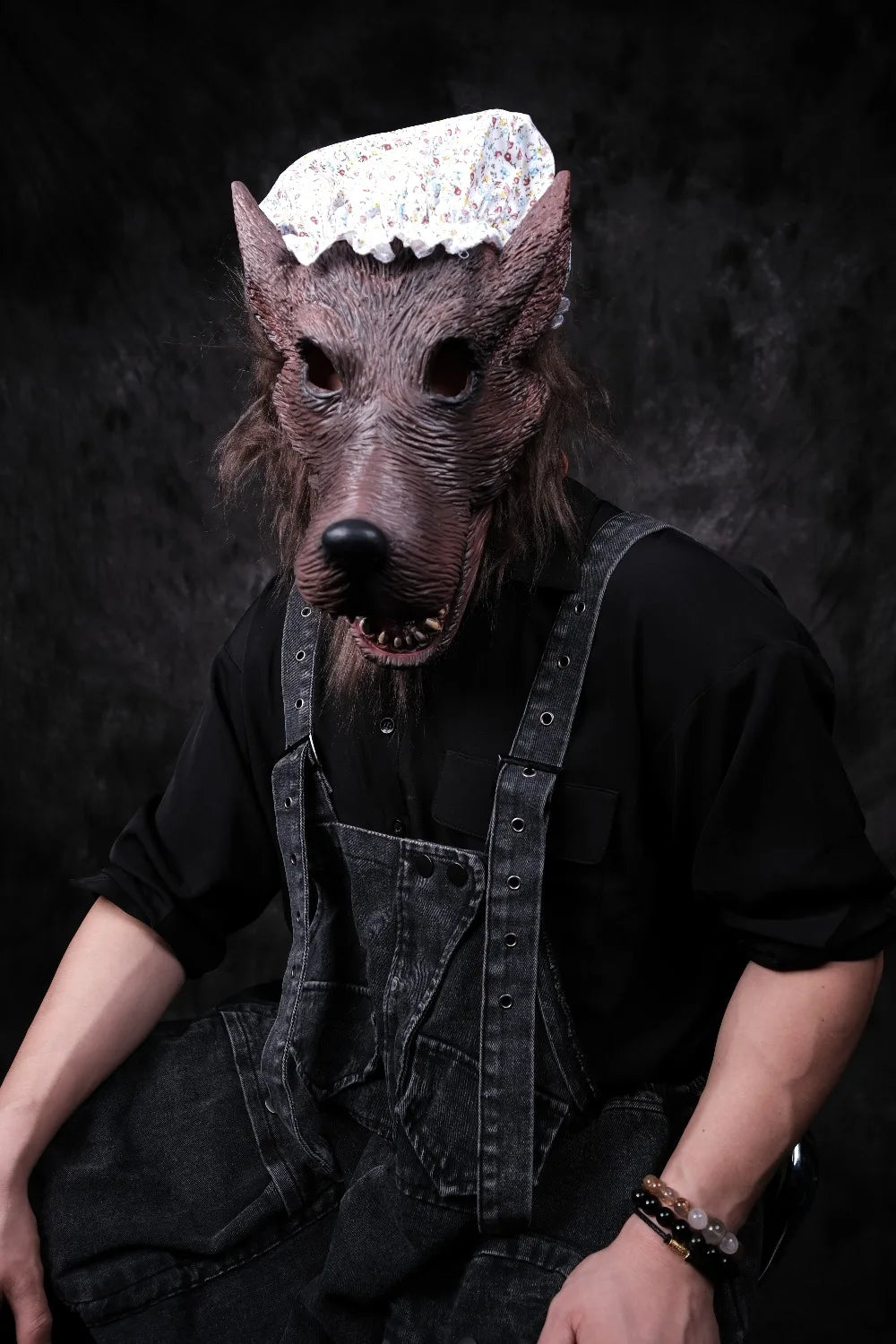 Granny Wolf Costume Mask shown on a seated wearer in black clothing and denim overalls