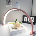 Half Moon Nail Lamp in pink on a white desk, framing a cream rose resting on stacked books.