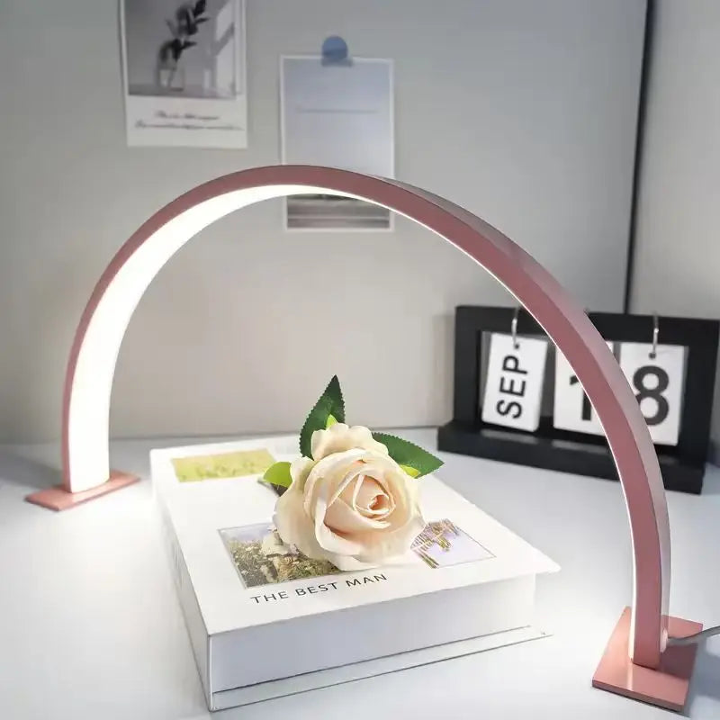 Half Moon Nail Lamp in pink on a white desk, framing a cream rose resting on stacked books.