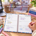 An open spiral-bound 2026/2027 weekly planner shown on a desk with a hand writing in a pink pen. The pages are floral with white writing space and date headers from August 26 to September 3.