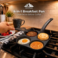 A 4-in-1 breakfast pan on a stovetop in a kitchen setting, cooking a sausage patty, biscuit, fried egg, and pancake. A hand is visible holding a spatula near the pan, with a mug and kitchen items in the background.