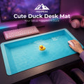 A desk mat shaped like a shallow pool of water with a small yellow duck floating in the center, shown on a dark desk with a keyboard, mouse, headphones, and a visible hand using the mouse. The text 'HIGHPEAK Cute Duck Desk Mat' appears at the top.