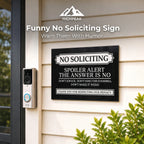 A black rectangular humorous 'No Soliciting' sign mounted on a beige house exterior door, next to a Ring doorbell camera. The sign features white text and decorative borders.