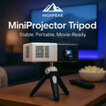 HighPeak MiniProjector Tripod in black supporting a compact projector on a wooden table in a cozy home theater setup