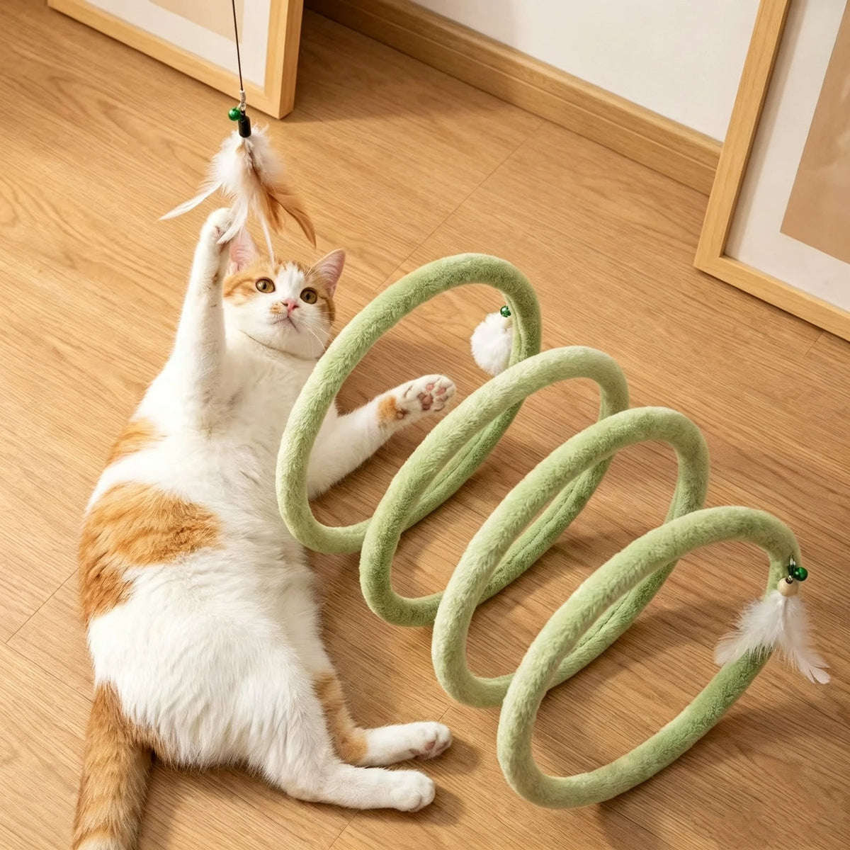 Orange and white cat reaching for a held feather beside a green Plush Cat Spiral Toy on a wood floor