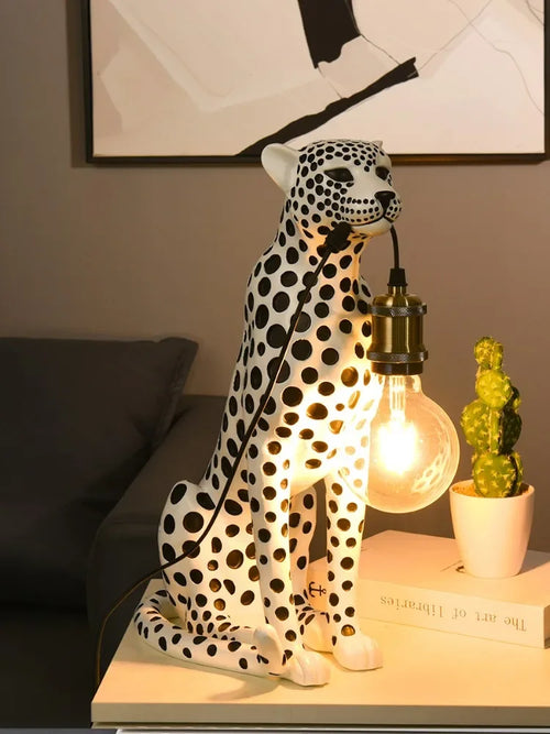 White Spotted Cheetah Premium Lamp holding a lit globe bulb by its cord on a side table beside a cactus and books.