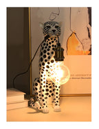 White Spotted Cheetah Premium Lamp viewed from the front with a glowing hanging bulb, books and framed artwork.
