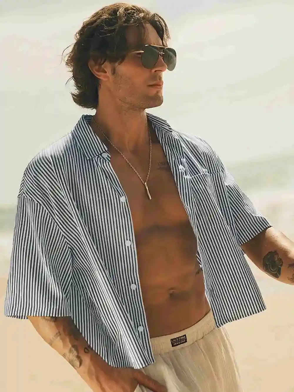 Men’s Striped Beach Shirt by HighPeak in blue and white stripes worn open on beach with sunglasses and light shorts