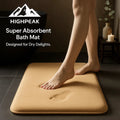 HighPeak Super Absorbent Bath Mat in tan with anti-slip backing, soft texture, shown with bare feet in dark bathroom setting