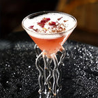 Jellyfish Cocktail Glass holding a pink drink topped with white foam and petals on a dark textured surface.