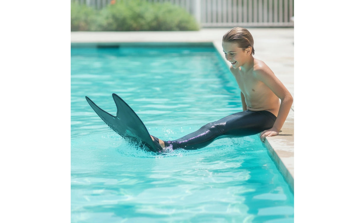 Kids Mermaid Swim Fin in a wide poolside scene showing a seated child splashing with the black fin.