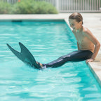 Kids Mermaid Swim Fin in black worn by a child splashing at the edge of a swimming pool.