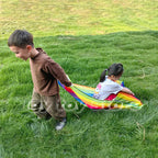 Kids Rainbow Pull Game Mat held by a walking boy with a girl seated in the striped pocket on a lawn
