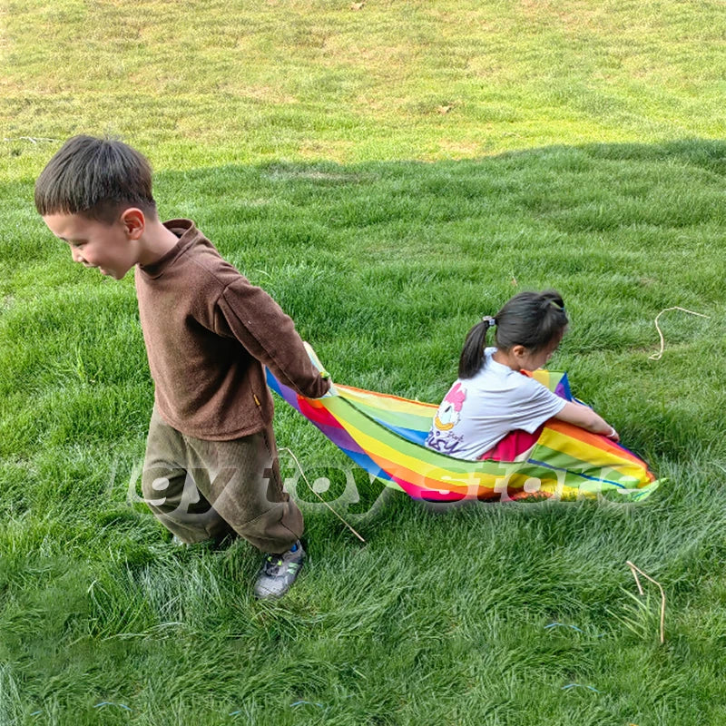 Kids Rainbow Pull Game Mat held by a walking boy with a girl seated in the striped pocket on a lawn