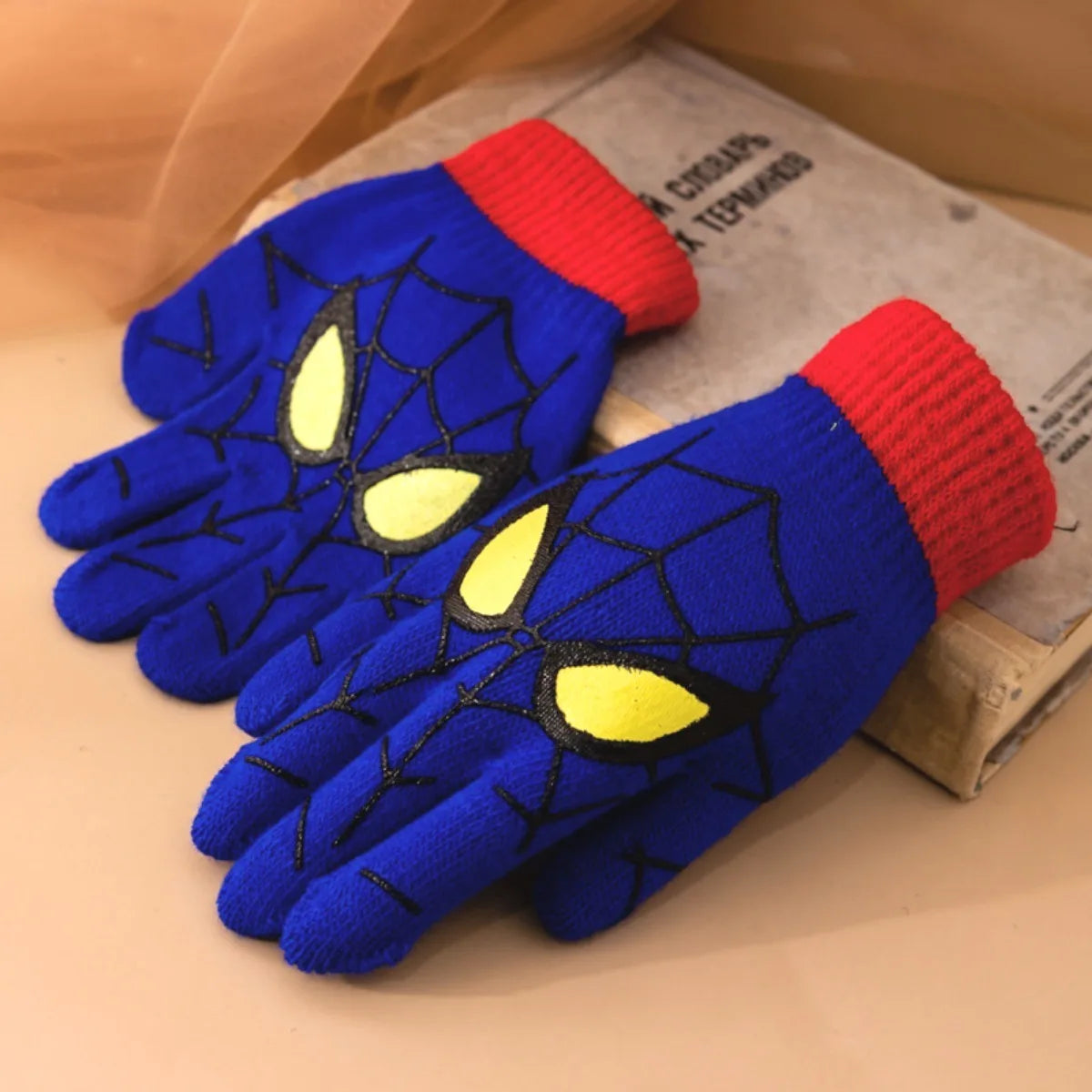 Blue kids spider web gloves with red ribbed cuffs arranged beside a book on a warm-toned surface.