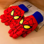 Red kids spider web gloves with blue ribbed cuffs arranged beside a book on a warm-toned surface.