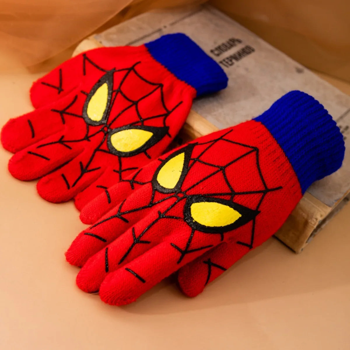 Red kids spider web gloves with blue ribbed cuffs arranged beside a book on a warm-toned surface.
