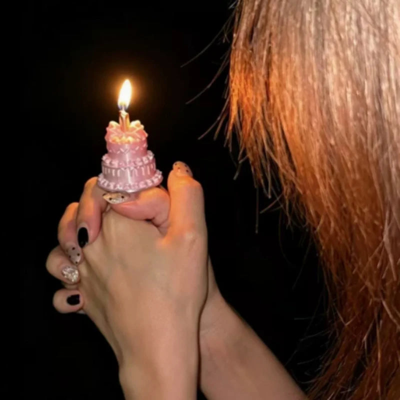 Mini Birthday Cake Ring in pink worn on a hand, with a lit candle against a dark party background.