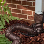 Retractable Downspout Extension in brown connected to a white downspout beside a brick wall and mulch