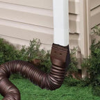 Retractable Downspout Extension in brown attached to a white downspout beside light siding and shrubs
