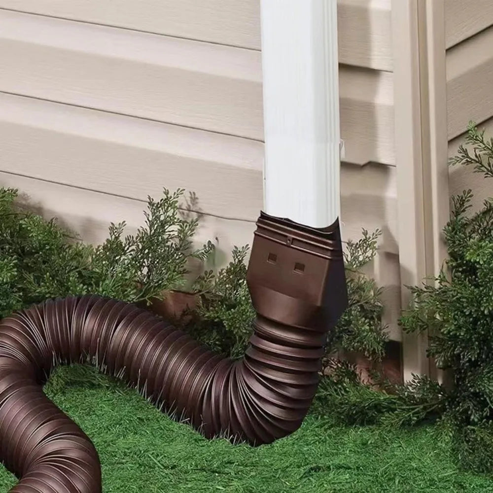 Retractable Downspout Extension in brown attached to a white downspout beside light siding and shrubs