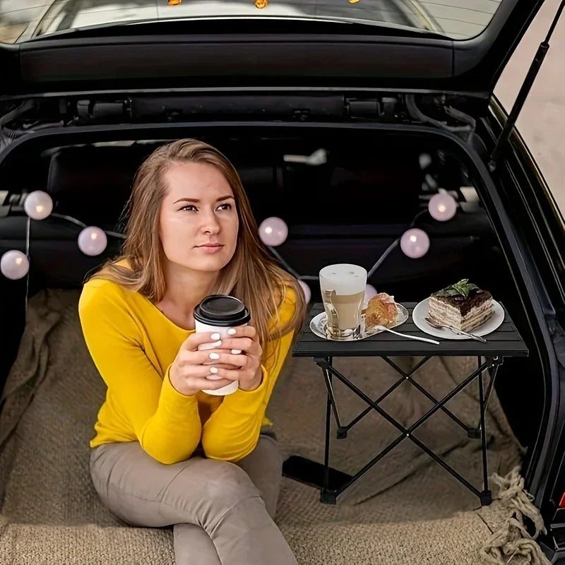 Roll-Up Camping Table used for a road-trip snack break in a car trunk decorated with string lights.