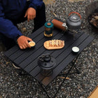 Roll-Up Camping Table overhead campsite view with a camper reaching for a cup beside snacks and a kettle.